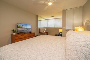 Un dormitorio con cama, televisor sobre una cómoda de madera, ventilador de techo y ventana con persianas.