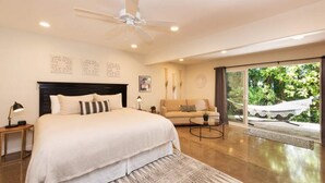 Une chambre moderne avec un grand lit, un canapé et une table basse. On y trouve également un ventilateur de plafond, une table de chevet et une fenêtre donnant sur un paysage verdoyant.