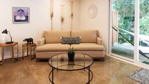 Un salon avec un canapé beige, une table basse en verre, une petite table d’appoint avec une lampe et un tableau encadré accroché au mur.