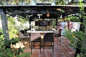 An outdoor patio with a brick floor, a wooden table, wicker chairs, and a TV mounted on a wooden structure. - The Seahorse Suite.  Welcoming guests back to Ft Myers Beach!  2 bedroom, 2 bath (Fort Myers Beach)