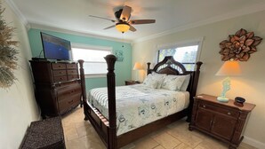 A four-poster bed with a headboard, a nightstand with a lamp, a dresser, and a decorative wall piece. - The Seahorse Suite.  Welcoming guests back to Ft Myers Beach!  2 bedroom, 2 bath (Fort Myers Beach)