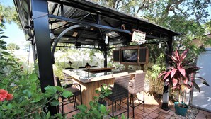 An outdoor bar area with a grill, seating, and a TV screen. - The Seahorse Suite.  Welcoming guests back to Ft Myers Beach!  2 bedroom, 2 bath (Fort Myers Beach)