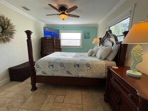 A bedroom with a four-poster bed, a ceiling fan, a window with blinds, a dresser, and a nightstand with a lamp.