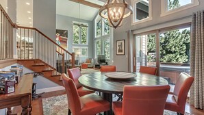 Uma sala de estar espaçosa com uma mesa redonda de madeira e poltronas de couro vermelho, escada com guarda-corpo de madeira e janelas amplas com vista para o exterior.