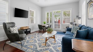 Un salon comprenant un canapé, un fauteuil, une table basse et un téléviseur. On y trouve également une table à manger avec des chaises, ainsi qu’une fenêtre avec des stores.