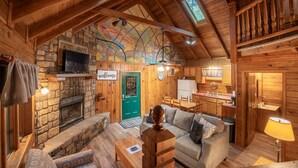 L’intérieur d’un chalet confortable, avec une cheminée en pierre, des poutres en bois au plafond, une porte verte et un coin cuisine.