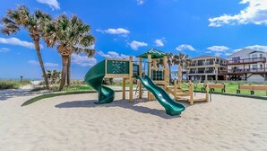 A beachside playground with a slide and climbing structure, surrounded by palm trees and a clear sky.