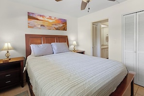 A bedroom with a bed, two bedside lamps, a nightstand, a wooden headboard, a painting on the wall, a closet, and a door leading to a bathroom. - Ocean View Condo with Large Lanai (Kailua)