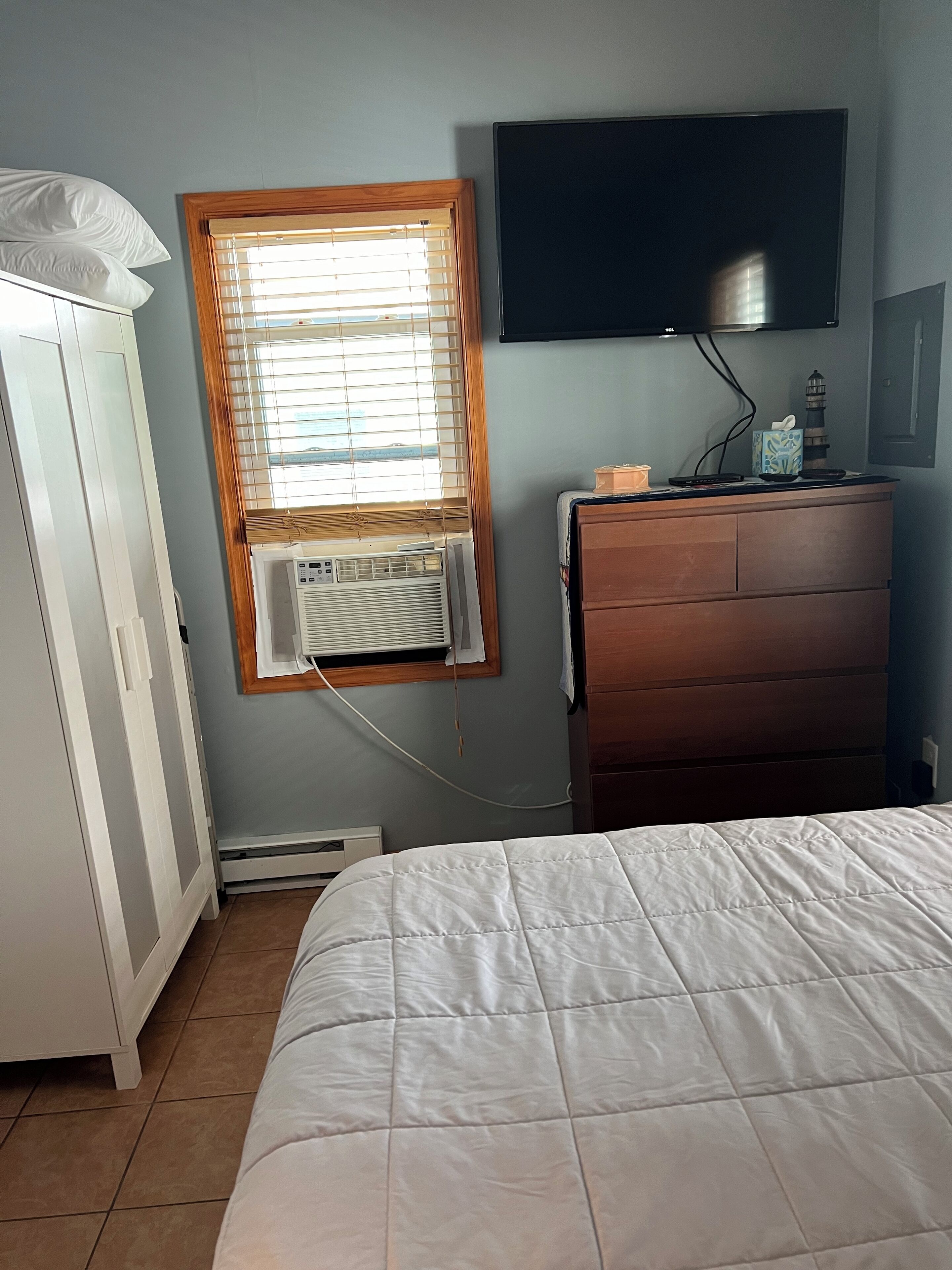 A bedroom with a bed, a dresser, a television mounted on the wall, and an air conditioning unit.