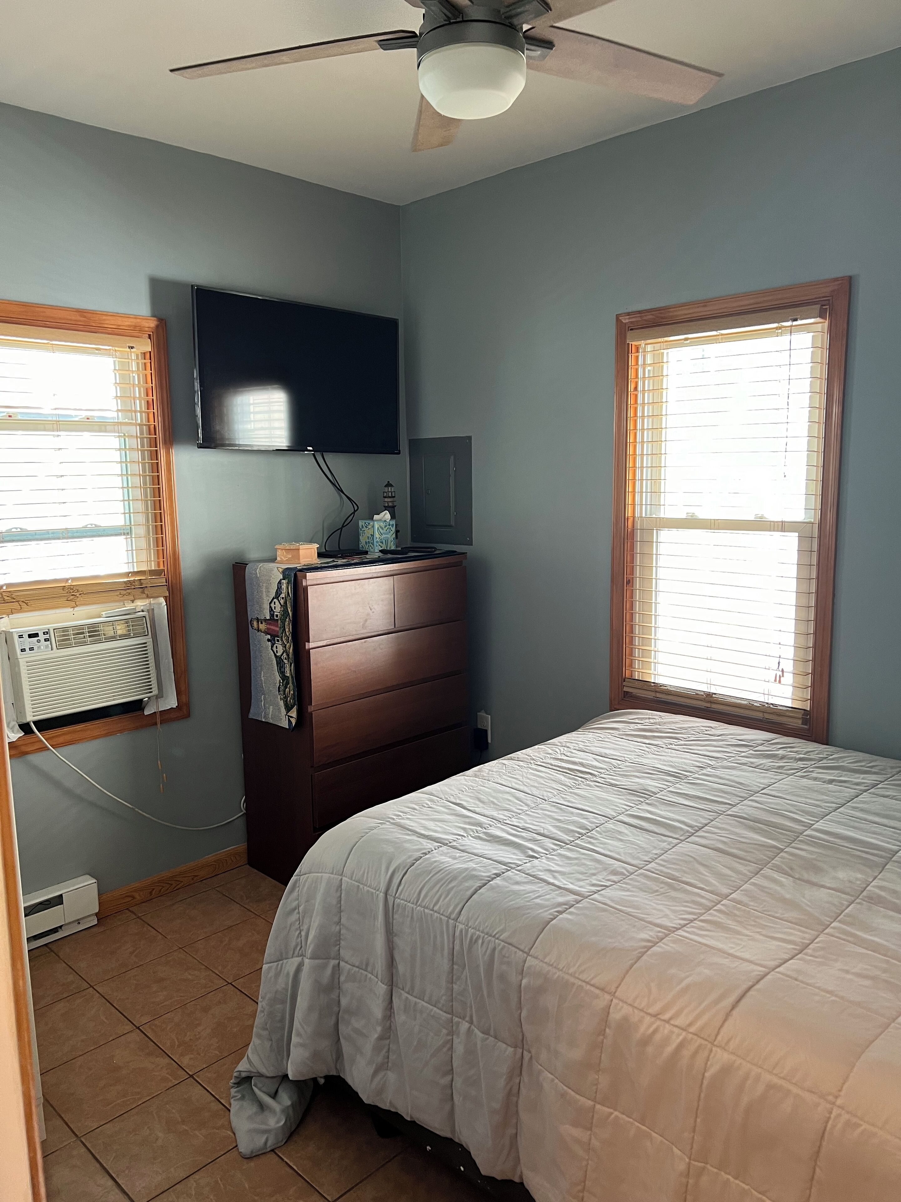 Un dormitorio con cama, cómoda, ventilador de techo, televisor montado en la pared y ventana con persianas.