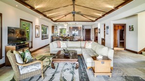 A spacious living room with a high ceiling, a large sofa, a television, and a dining area with a kitchen in the background. - Serene 3BR Puako Estate P10D – Beachfront Access & Tranquil Living (Kamuela)