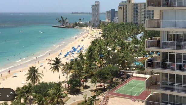 A beach with palm trees, a tennis court, and high-rise buildings in the background. - FLASH SALE:  Beautiful Beach Front Location (Carolina, Isla Verde)