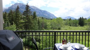 A balcony with a table set for two, overlooking a mountainous landscape with dense forests. - 🍁Mountain Views, Pools, Fitness, Restaurant & MORE, we're accepting bookings! (Canmore)