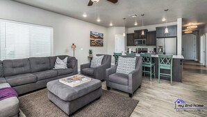 A modern living room with a gray sofa, two armchairs, and a coffee table. There is a ceiling fan, recessed lighting, and a kitchen in the background. - Estancia S304 Poolside Getaway Pool (St. George)