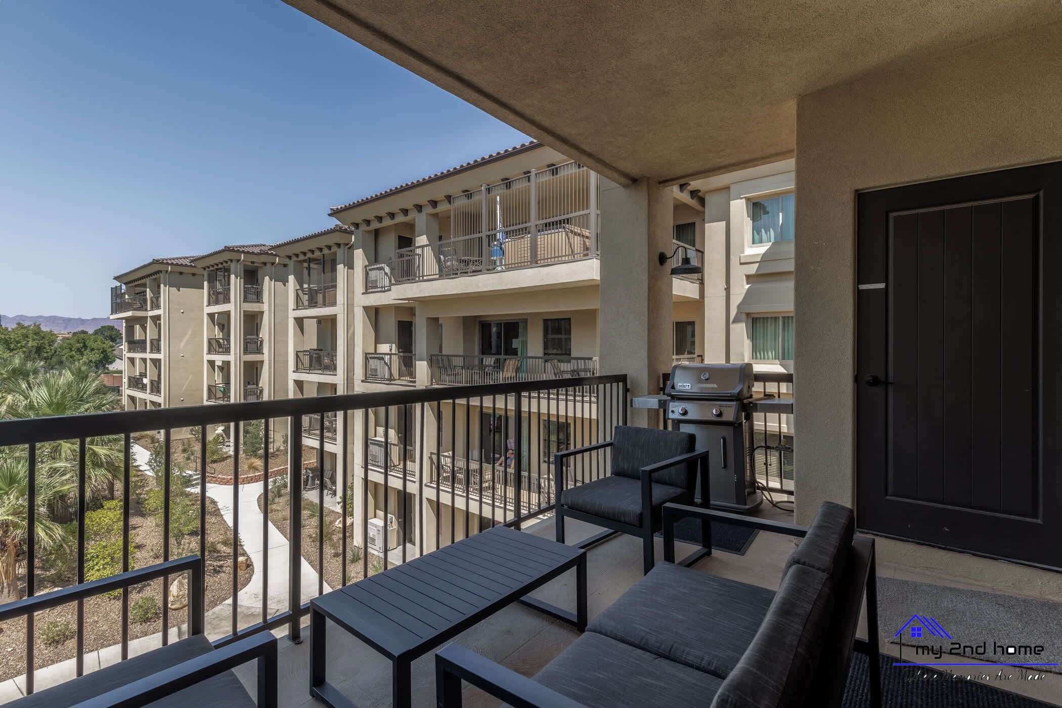Een balkon met buitenmeubilair, een barbecue en uitzicht op andere gebouwen en groen.