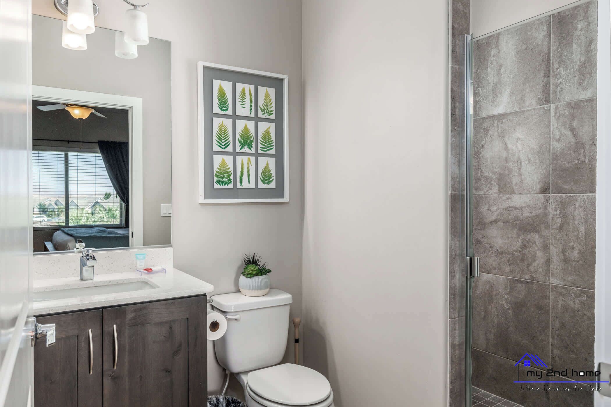 Une salle de bain moderne avec des toilettes, un lavabo et une douche. Un tableau encadré représentant des feuilles est accroché au mur.