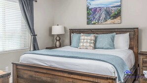 A bedroom with a wooden bed, a nightstand with a lamp, a window with blinds, and a painting of a canyon on the wall. - Estancia S304 Poolside Getaway Pool (St. George)