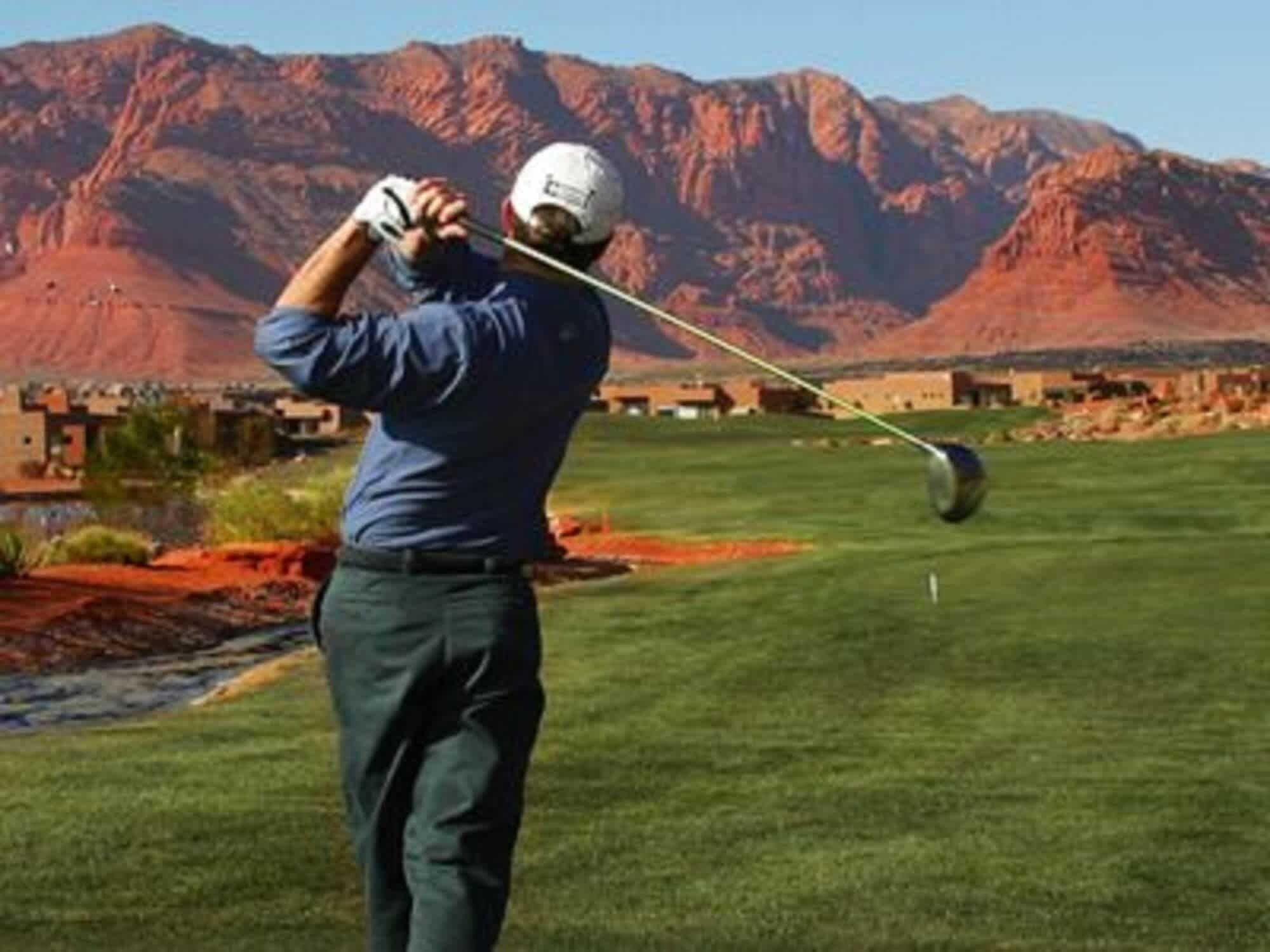 A golfer in a blue shirt and white helmet preparing to swing at a golf course with a mountainous backdrop.