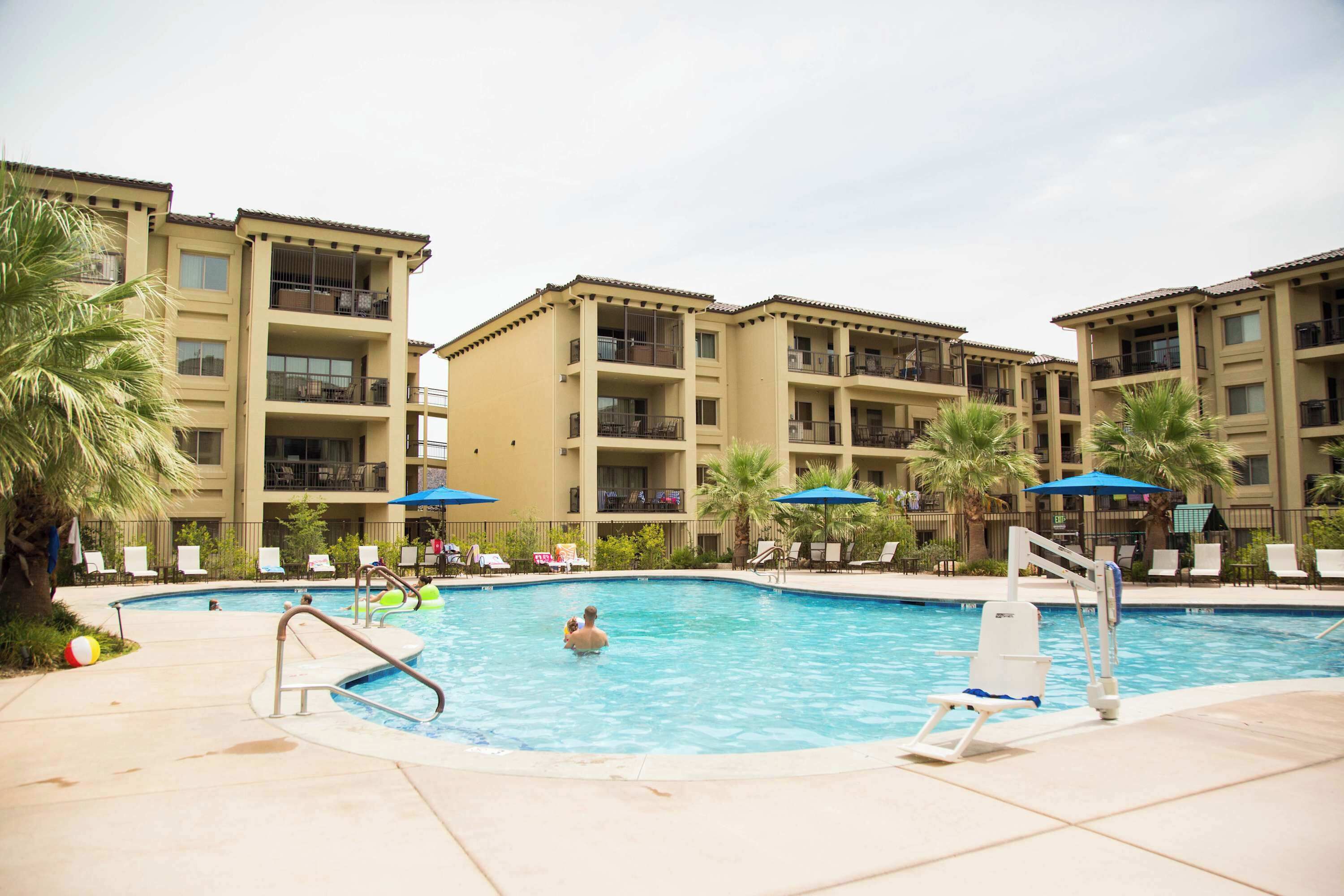 A swimming pool with lounge chairs and umbrellas, surrounded by multi-story buildings with balconies.