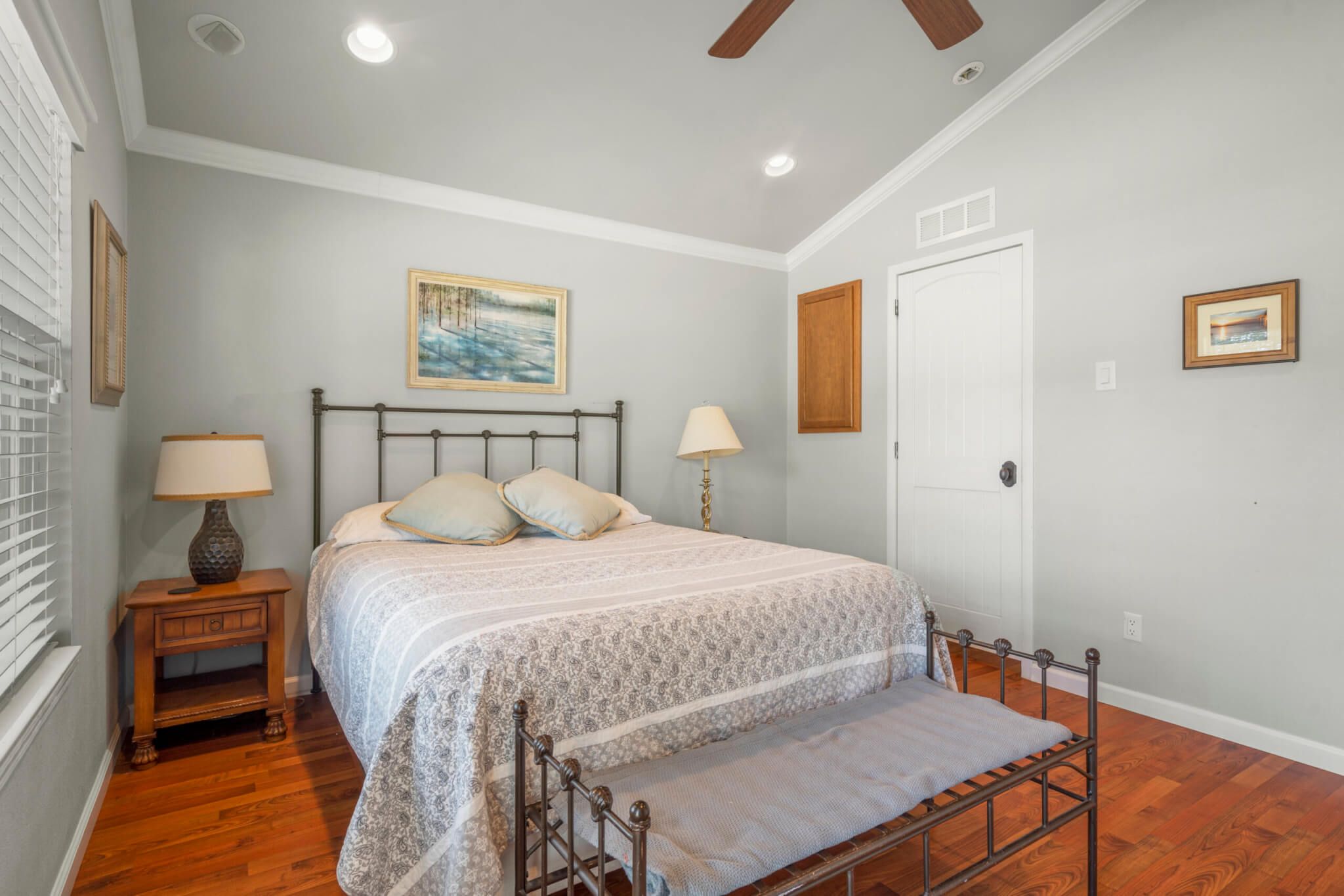 A bedroom with a bed, bedside tables, a bench, a ceiling fan, and a painting on the wall.