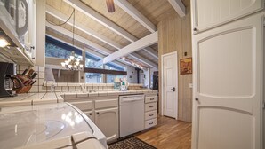 Een keuken met houten balken, witte kasten, een vaatwasser en een raam met uitzicht op een woning.
