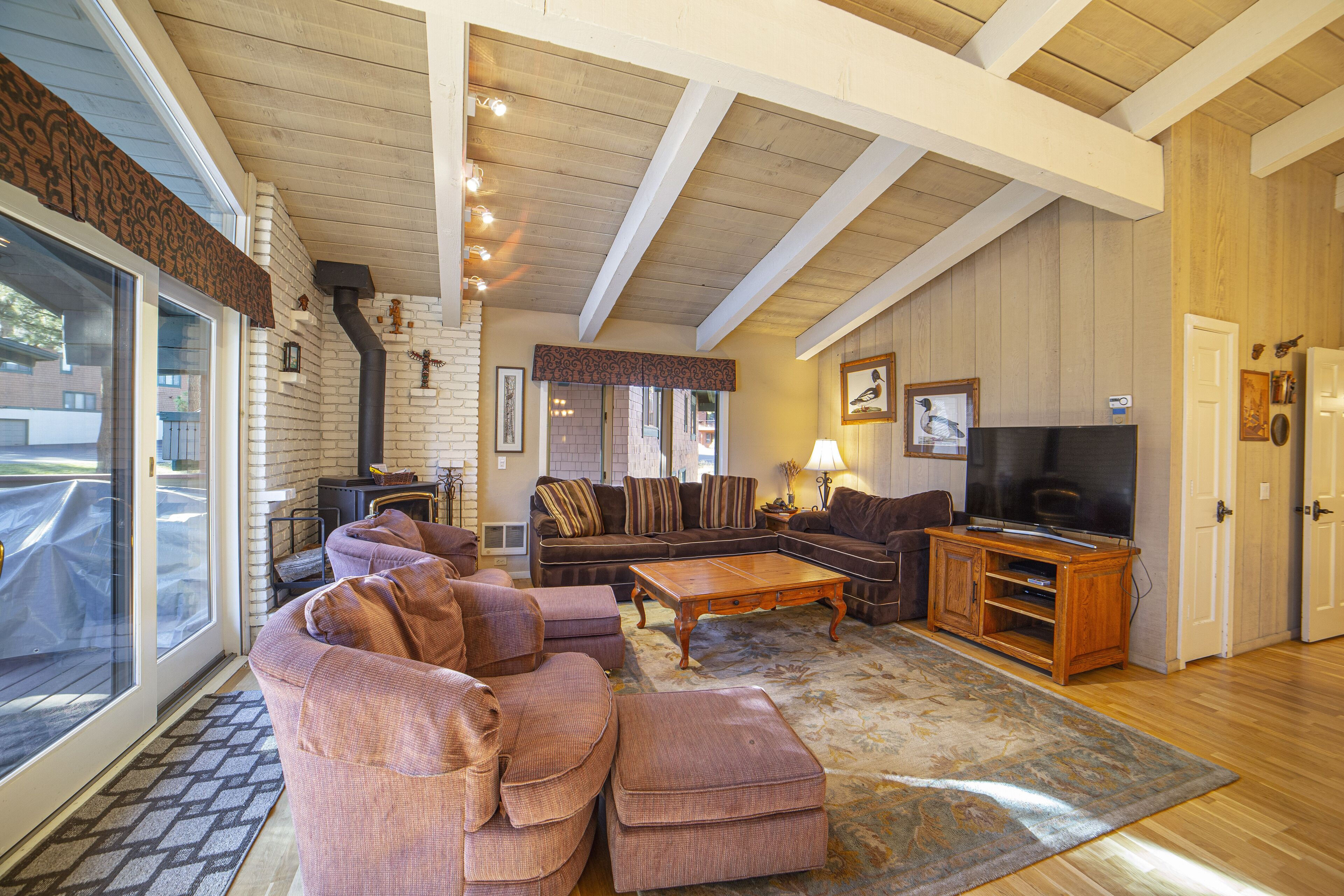 A spacious living room with a large sectional sofa, a coffee table, and a television. There is a fireplace, a large window, and a door leading outside.