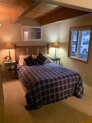 Un dormitorio con una cama, dos mesitas de noche con lámparas, una ventana con vista a un coche, y un cuadro enmarcado en la pared.