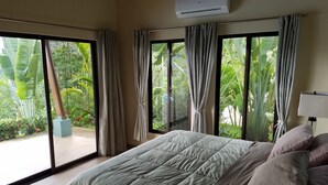 A bedroom with a bed, a window with curtains, an air conditioning unit, and a view of greenery outside. - Luxury Mountain Home with Ocean and Rain Forest Views.  Newly tiled pool! (Chontales)