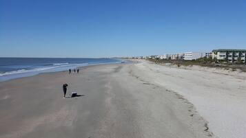 Una playa extensa con cielo despejado, algunas personas caminando y edificios al fondo.
