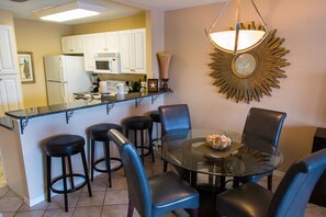 A dining area with a glass table, black chairs, and a bar counter with stools. - Lakefront 230D - Next to the pool and steps from the beach and Whale's Tail! (Miramar Beach)