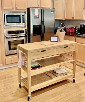 Eine Küche mit Holzausstattung, einem Edelstahlkühlschrank, einer Mikrowelle und einem Backofen. Ein Holzwagen mit Schubladen und Schneidebrett steht ebenfalls bereit.
