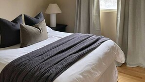Een net opgemaakt bed met kussens, een bedlampje en een raam met gordijnen.
