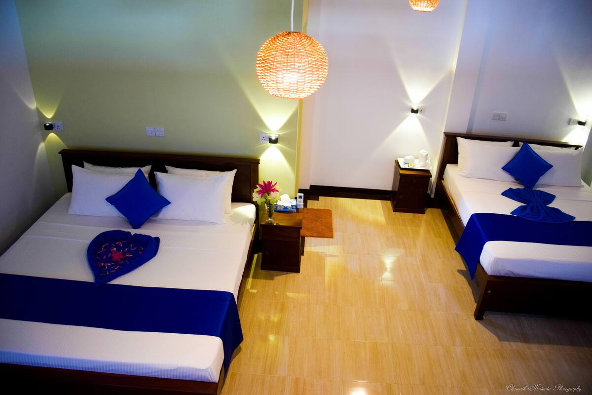 A hotel room with two beds, each with a blue bedspread and pillows, a bedside table with a vase of flowers, and a hanging light fixture.