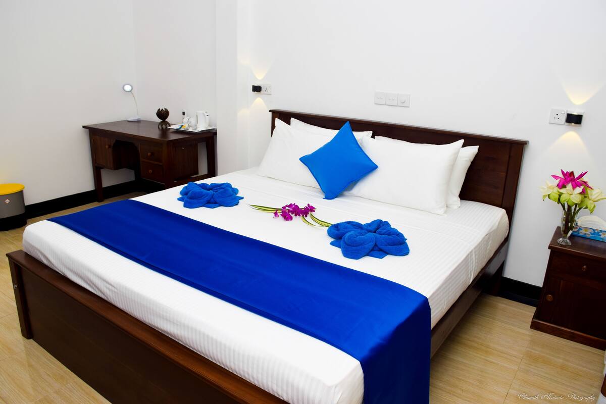 A neatly made bed with a blue bedspread, white pillows, and a blue accent pillow. There is a wooden nightstand with a lamp and a vase with flowers beside the bed.