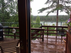 En træterrasse med gelænder, en stol og udsigt til skov og flod.