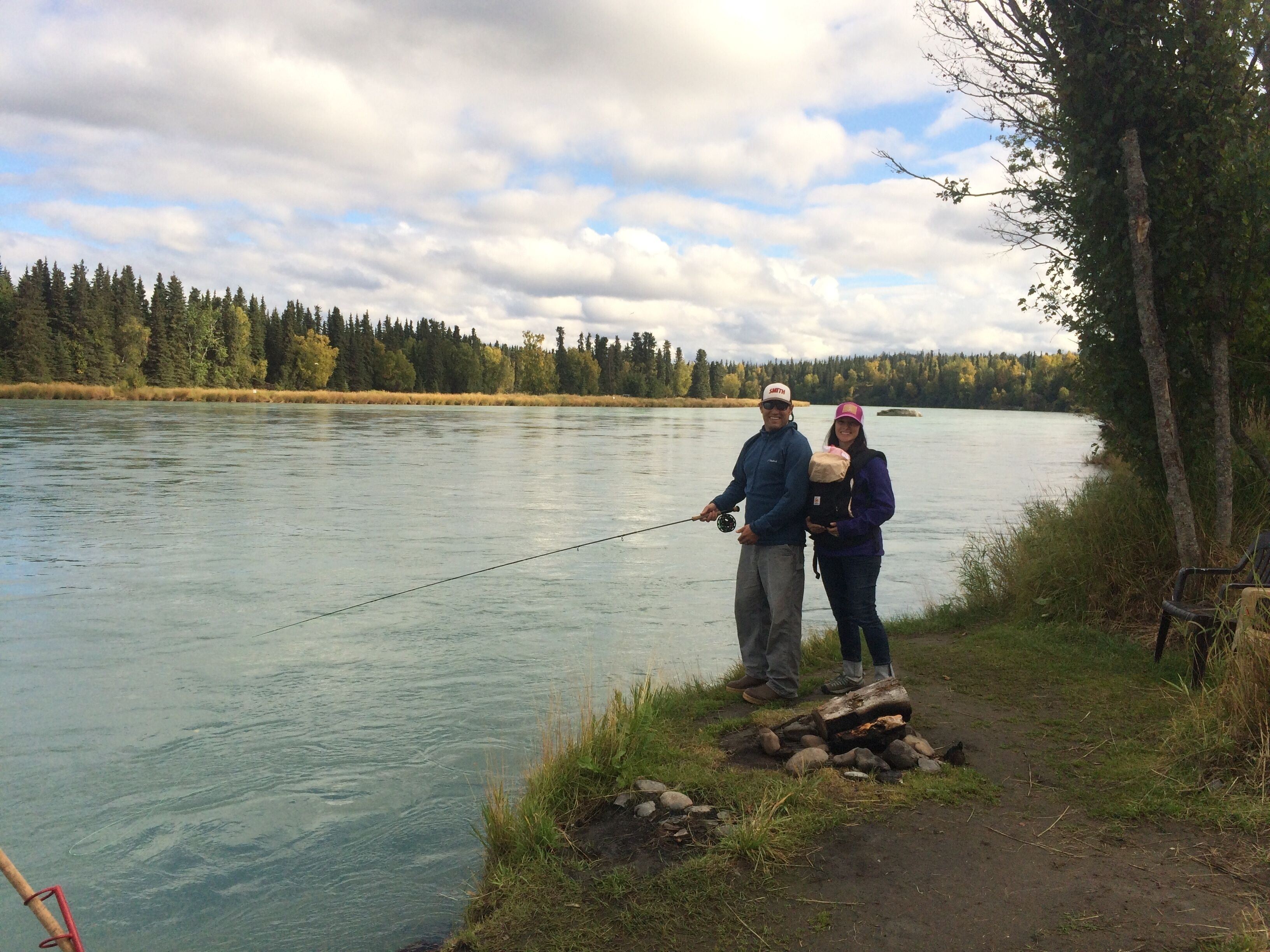 A couple fishing by a calm river with a forested background.