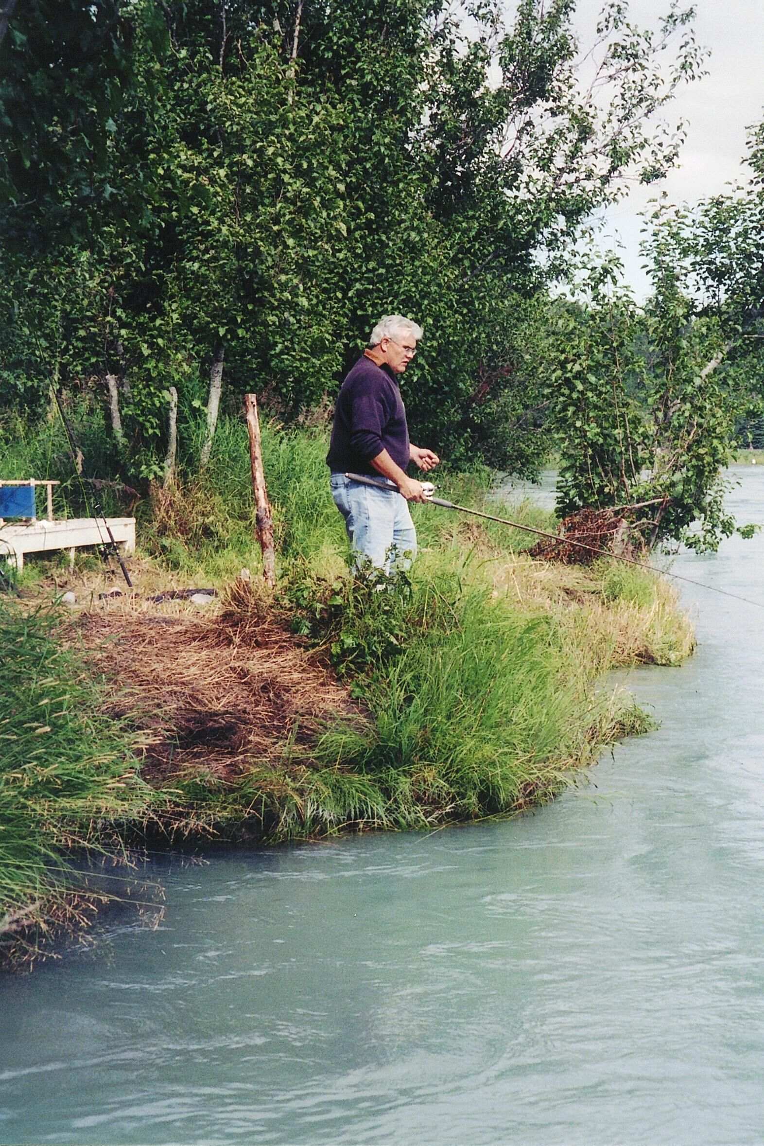 A man fishing by a calm river surrounded by dense greenery.