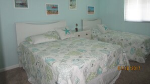 Une chambre avec deux lits, chacun doté d’une tête de lit blanche et d’une couvre-lit à motifs d’étoiles de mer. Trois tableaux encadrés ornent le mur, et une fenêtre avec des stores se trouve sur le côté droit.