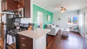 A modern kitchen with dark cabinets, a stove, and a microwave. There is a dining area with a white table and a gray sofa. The room has a green accent wall and a ceiling fan. - 1 Bedroom Condo steps from Ocean and Bay (San Diego)