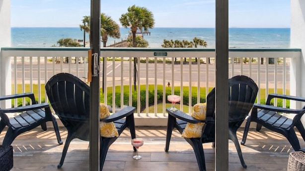 Ein Balkon mit zwei Stühlen und einem Tisch, mit Blick auf einen Strand und eine Straße.