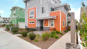 A modern two-story residential building with a grey and orange facade, a visible number '290', and a small front yard with plants and a black metal fence.