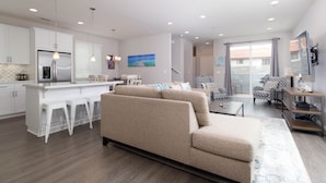 A modern living room with a beige sofa, a kitchen with white cabinets, and a dining area with bar stools.