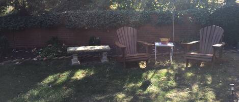 A garden with a stone bench and two Adirondack chairs, a small table, and a brick wall with climbing plants.