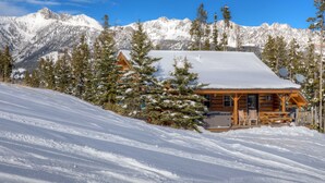 Una baita di legno con tetto innevato, circondata da alberi sempreverdi e montagne innevate sullo sfondo.