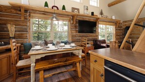 Interno di una baita rustica con un tavolo da pranzo in legno apparecchiato per quattro persone, una panca e un angolo cottura con lavastoviglie.