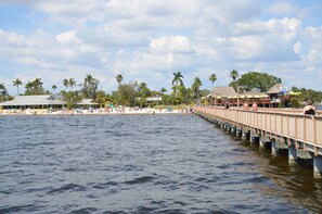 Un muelle que se adentra en el agua, con una playa y palmeras al fondo.