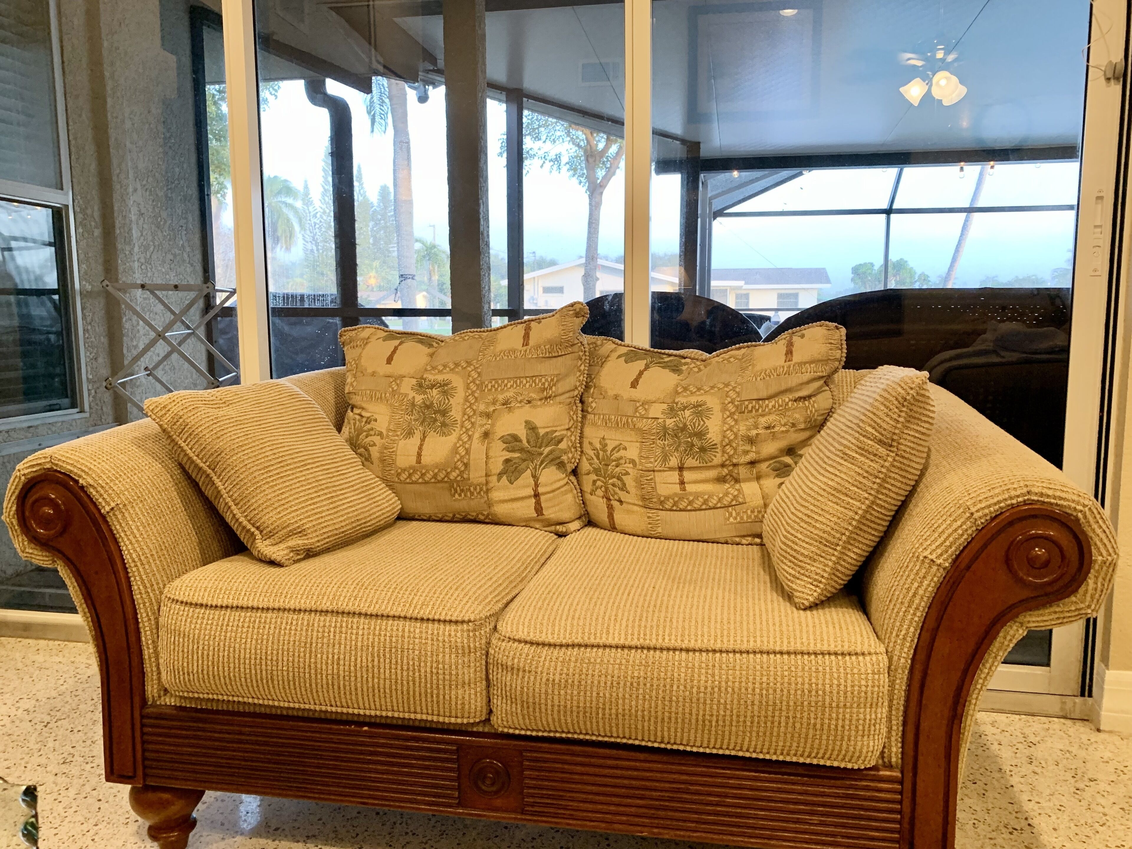 Ein beiges gewebtes Sofa mit floralen Kissen, vor einem Fenster mit Blick auf Palmen und Gebäude.