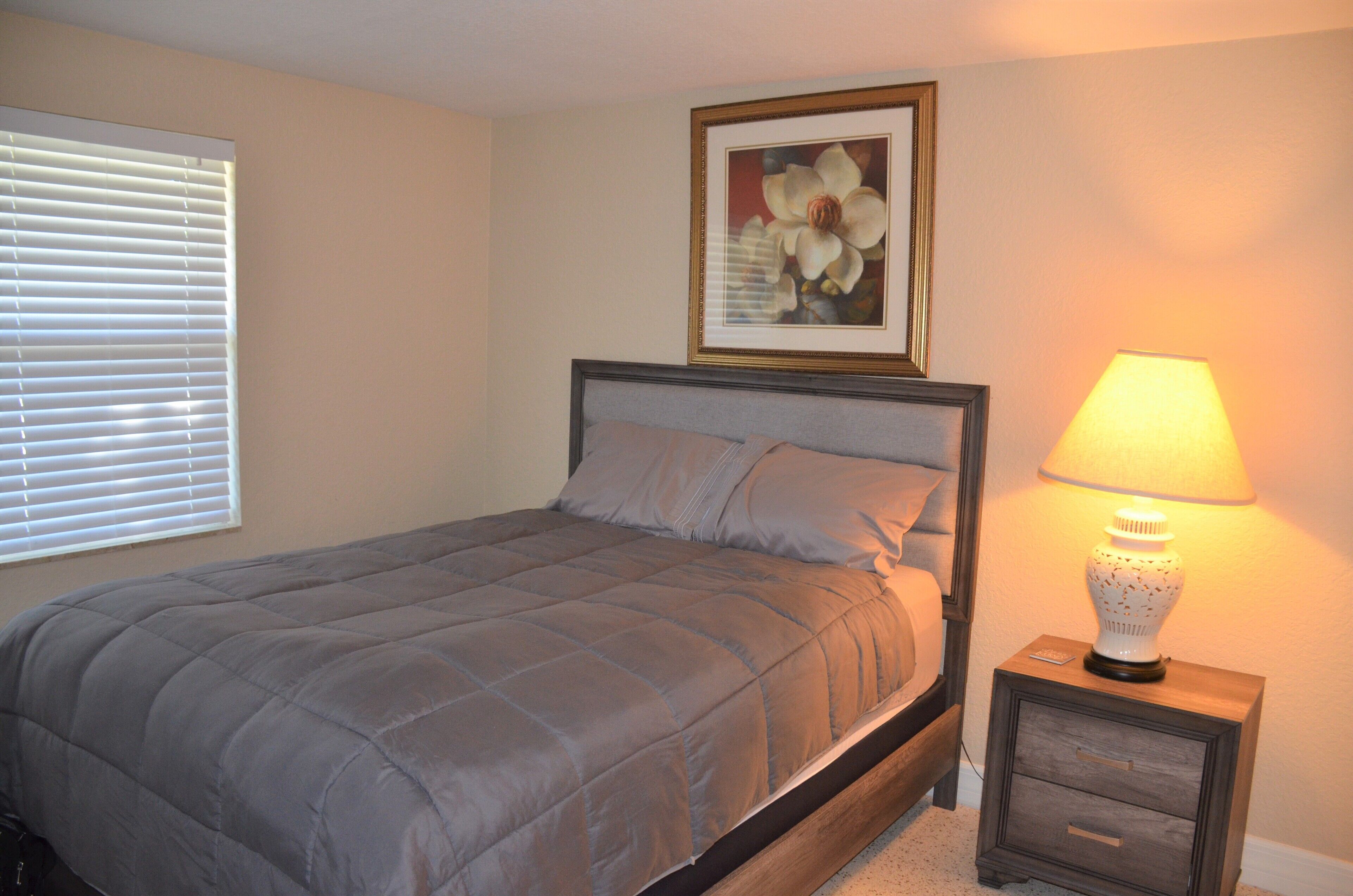 A bedroom with a bed, a nightstand with a lamp, a framed picture on the wall, and a window with blinds.