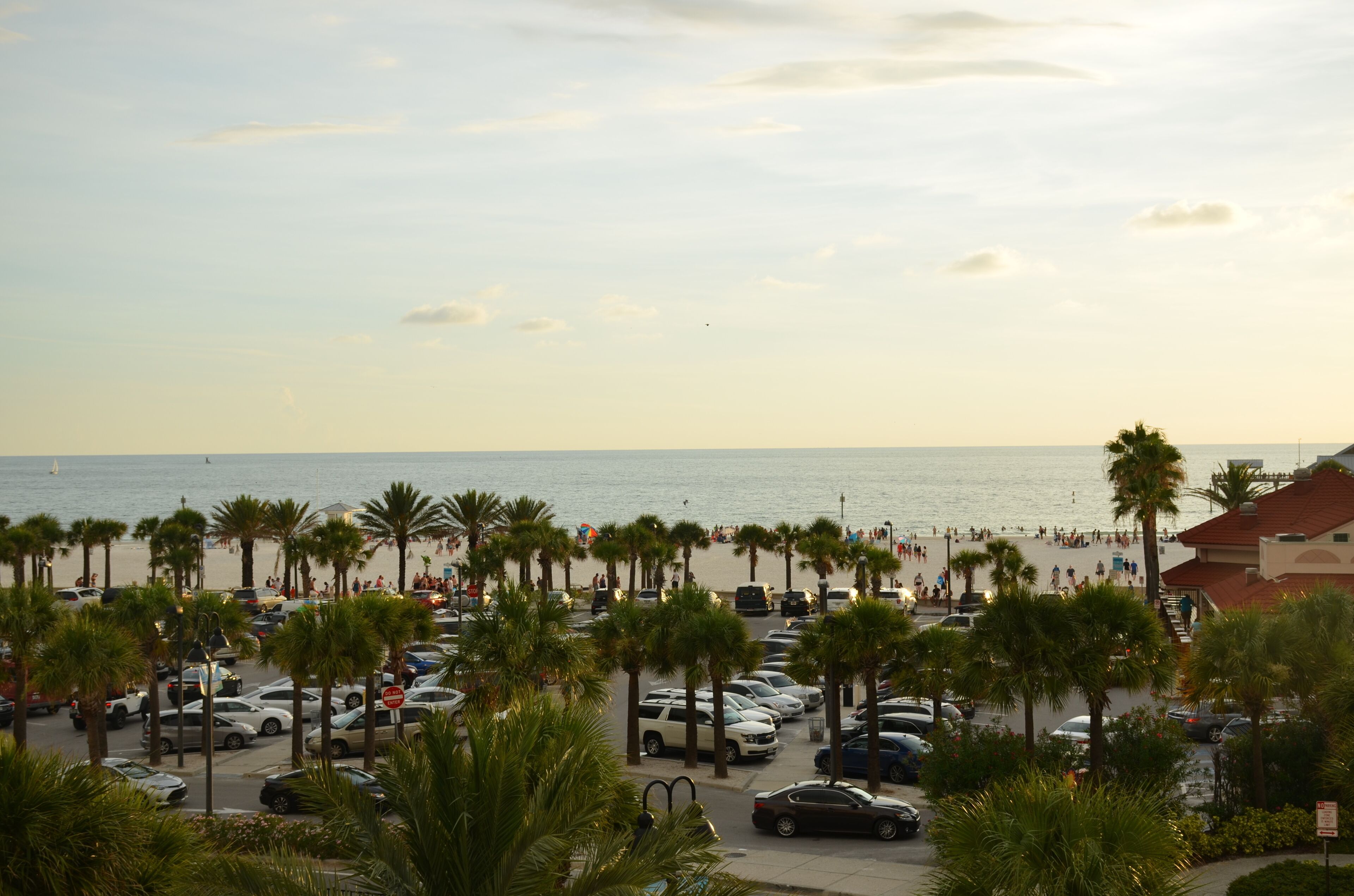 Un parcheggio vicino alla spiaggia con auto e palme, che conduce a una zona balneare affollata.