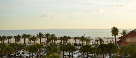 A beachside parking lot with cars and palm trees, leading to a crowded beach area.